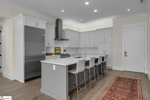 kitchen with wall chimney exhaust hood, stainless steel appliances, light hardwood / wood-style flooring, a breakfast bar, and a center island with sink