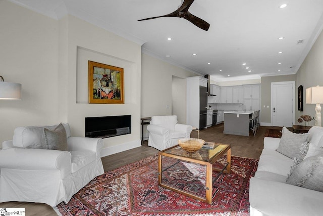 living room featuring ceiling fan, sink, ornamental molding, and dark wood-type flooring