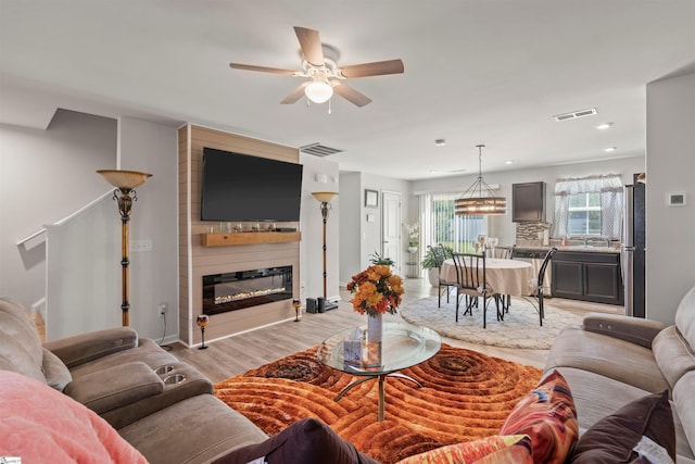 living room featuring a large fireplace, light wood-type flooring, and ceiling fan with notable chandelier