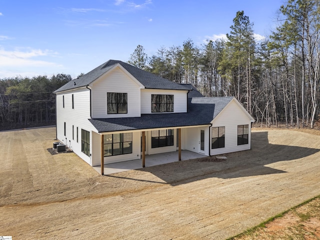 modern farmhouse with a shingled roof, cooling unit, a patio area, and a wooded view