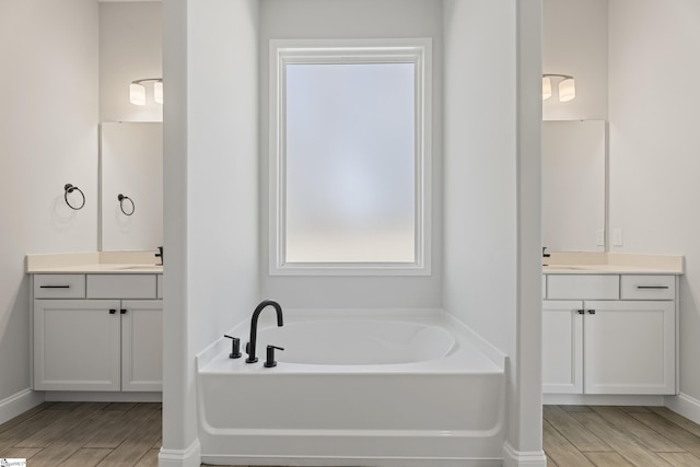 bathroom with baseboards, two vanities, a sink, wood finish floors, and a bath