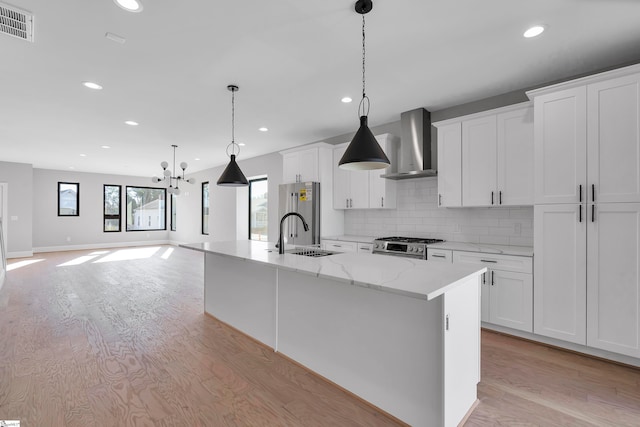 kitchen featuring appliances with stainless steel finishes, wall chimney range hood, pendant lighting, white cabinetry, and an island with sink