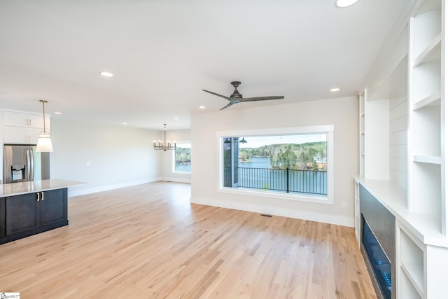 unfurnished living room with built in features, plenty of natural light, ceiling fan with notable chandelier, and light wood-type flooring