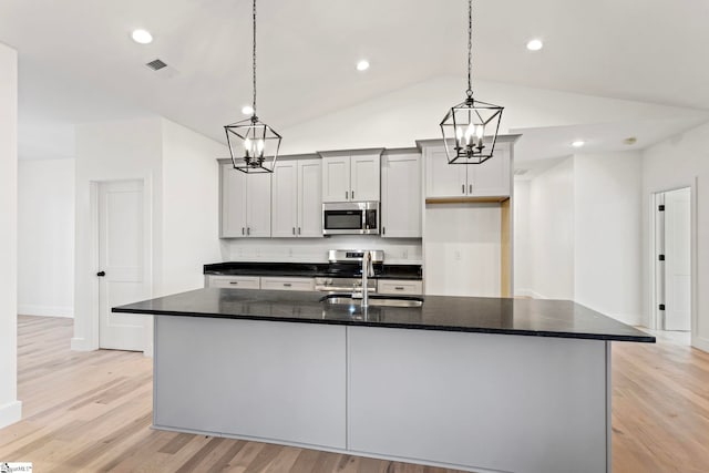 kitchen with appliances with stainless steel finishes, decorative light fixtures, dark stone countertops, light hardwood / wood-style floors, and a center island with sink