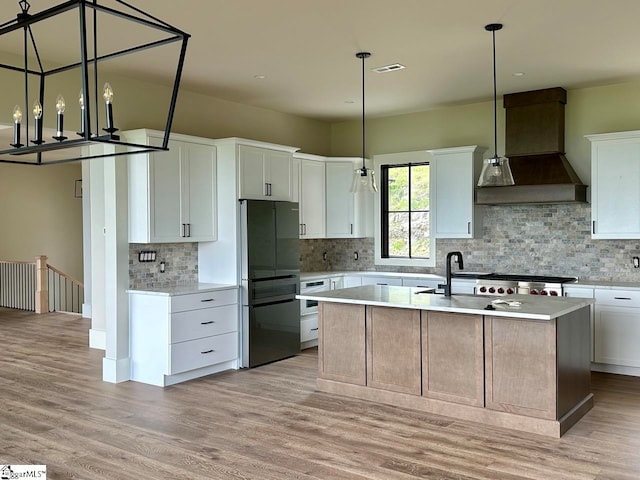 kitchen featuring light wood-type flooring, custom exhaust hood, white cabinets, hanging light fixtures, and an island with sink