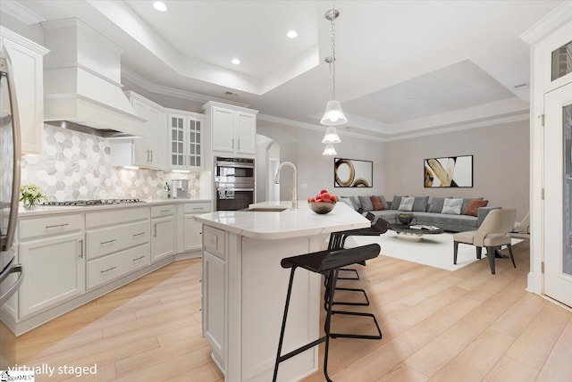 kitchen with custom exhaust hood, sink, backsplash, a raised ceiling, and an island with sink