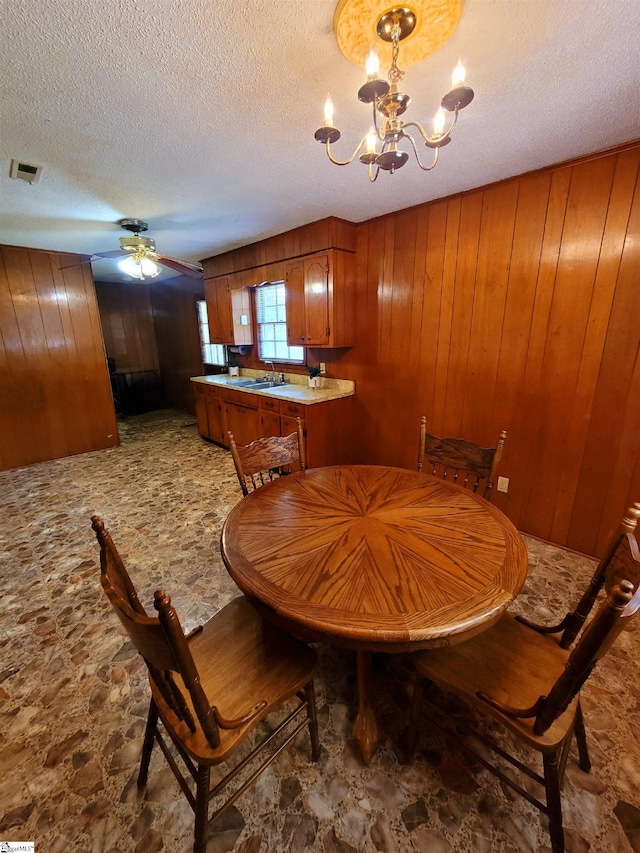 dining space with ceiling fan with notable chandelier, a textured ceiling, sink, and wood walls