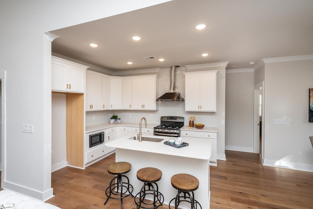 kitchen featuring black microwave, stainless steel gas stove, wall chimney exhaust hood, sink, and a center island with sink