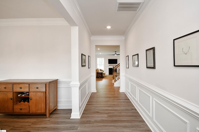 hall featuring wood finished floors, visible vents, a decorative wall, crown molding, and wainscoting