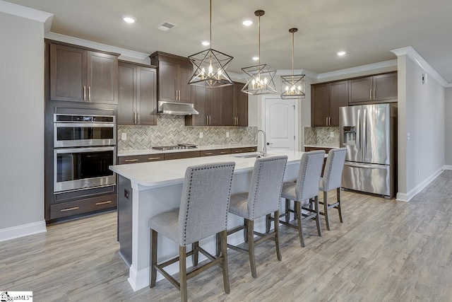 kitchen with a center island with sink, hanging light fixtures, appliances with stainless steel finishes, dark brown cabinets, and under cabinet range hood