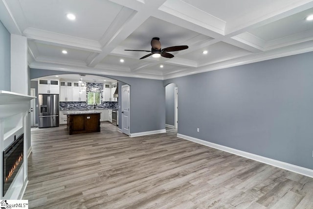 unfurnished living room with arched walkways, beam ceiling, a ceiling fan, coffered ceiling, and baseboards