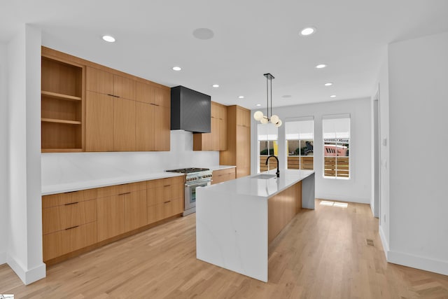 kitchen featuring stainless steel stove, wall chimney exhaust hood, an island with sink, sink, and decorative light fixtures