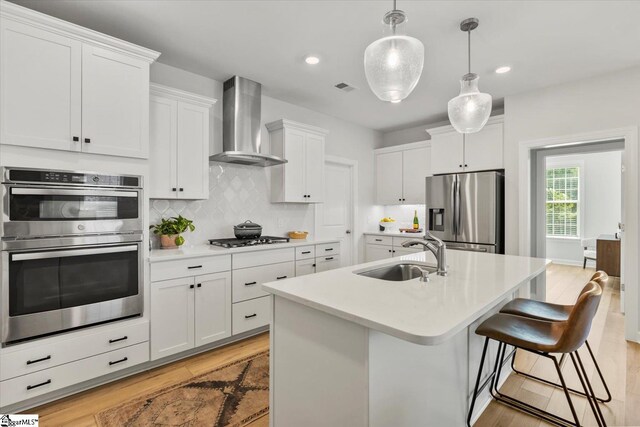 kitchen with white cabinetry, stainless steel appliances, a center island with sink, sink, and wall chimney range hood