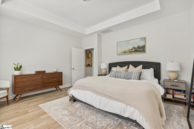 bedroom with light wood-type flooring, ensuite bath, and a raised ceiling