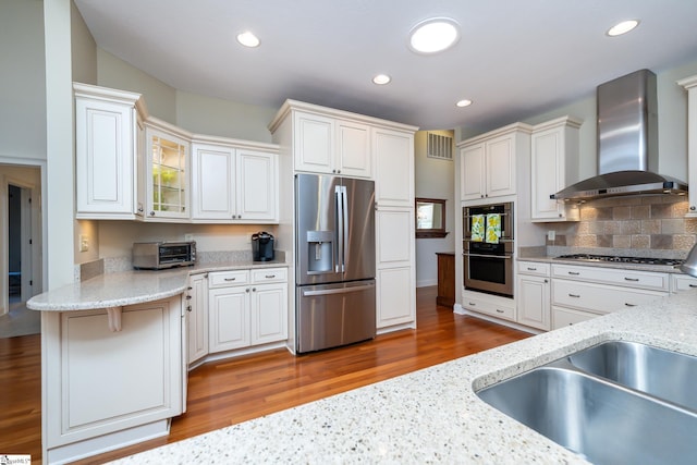 kitchen featuring white cabinetry, appliances with stainless steel finishes, wall chimney range hood, light stone countertops, and glass insert cabinets