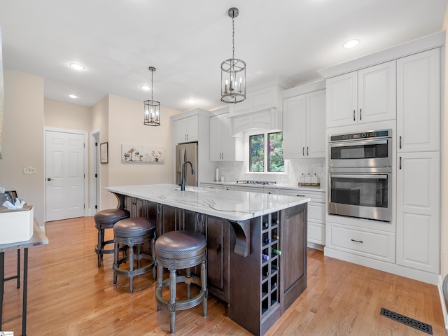 kitchen with a kitchen island with sink, stainless steel appliances, white cabinetry, a chandelier, and light wood-type flooring