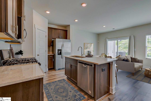 kitchen with light stone countertops, a kitchen island with sink, stainless steel appliances, sink, and light wood-type flooring