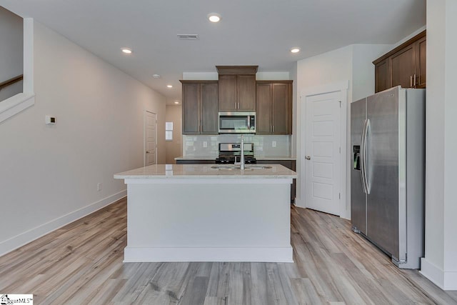 kitchen with a kitchen island with sink, light wood-type flooring, sink, light stone countertops, and appliances with stainless steel finishes