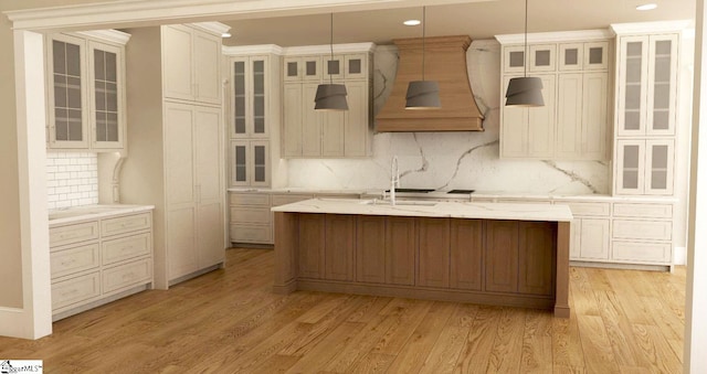 kitchen featuring light wood-type flooring, tasteful backsplash, decorative light fixtures, premium range hood, and a center island with sink