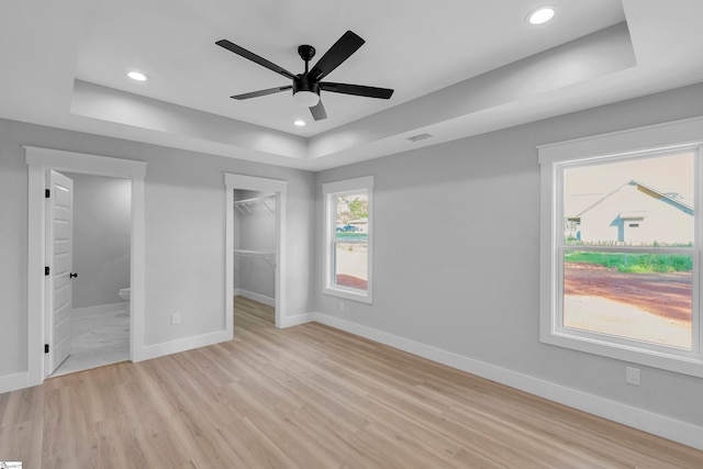 unfurnished bedroom featuring light hardwood / wood-style flooring, ensuite bath, ceiling fan, a raised ceiling, and a walk in closet