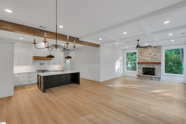 kitchen with ceiling fan with notable chandelier, decorative light fixtures, a stone fireplace, an island with sink, and white cabinets