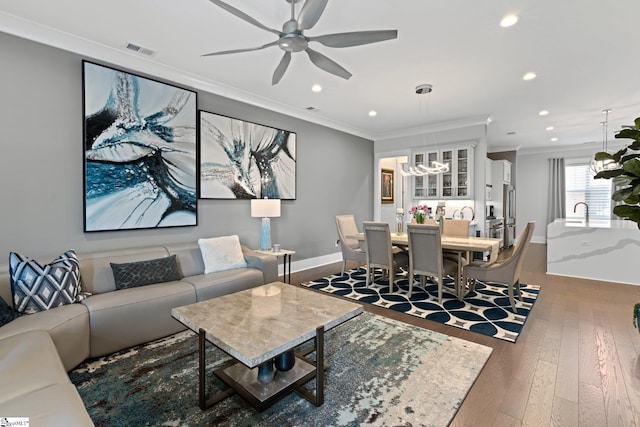 living room with dark wood-type flooring, ornamental molding, ceiling fan with notable chandelier, and sink