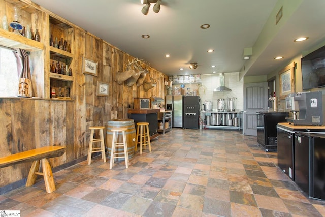 kitchen featuring wall chimney exhaust hood, wood walls, stainless steel fridge, and a kitchen breakfast bar