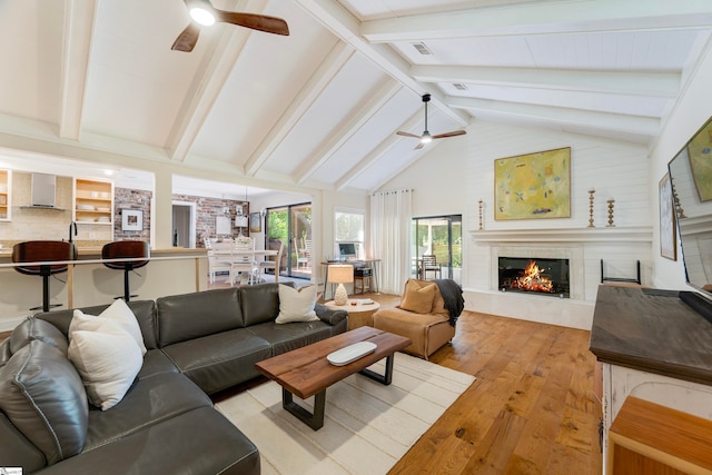 living room with ceiling fan, beam ceiling, light hardwood / wood-style flooring, high vaulted ceiling, and a fireplace