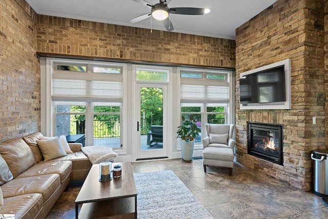 living room featuring ceiling fan, brick wall, crown molding, and a stone fireplace