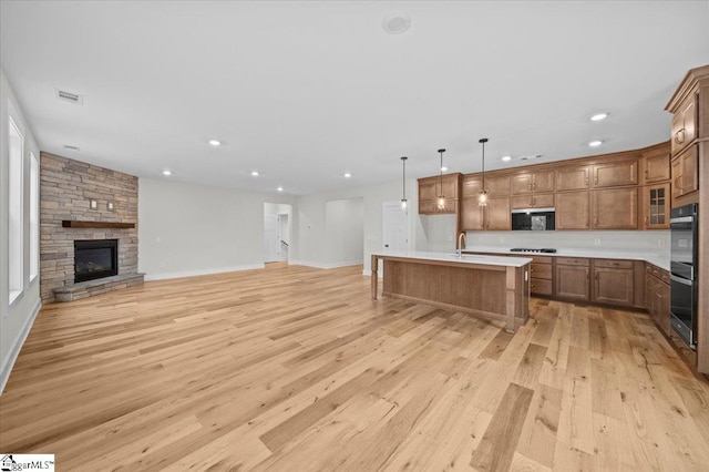 kitchen featuring a stone fireplace, double oven, an island with sink, decorative light fixtures, and light wood-type flooring