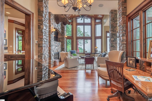 home office featuring wood-type flooring, a stone fireplace, ceiling fan with notable chandelier, and high vaulted ceiling