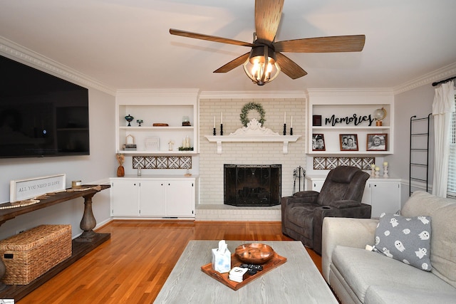 living room featuring ceiling fan, built in shelves, a fireplace, light wood-type flooring, and crown molding