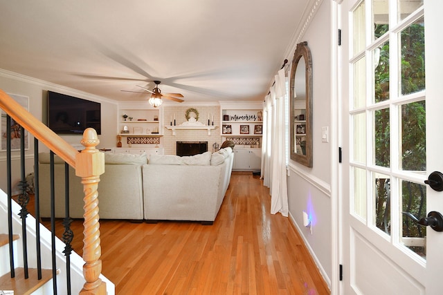 living room featuring built in shelves, ceiling fan, a fireplace, crown molding, and light hardwood / wood-style floors