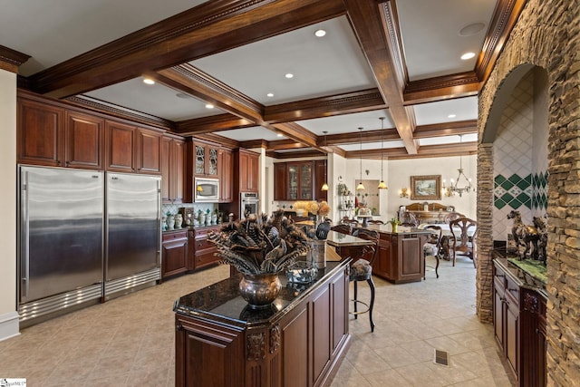 kitchen with a kitchen island, coffered ceiling, built in appliances, dark stone countertops, and a kitchen bar