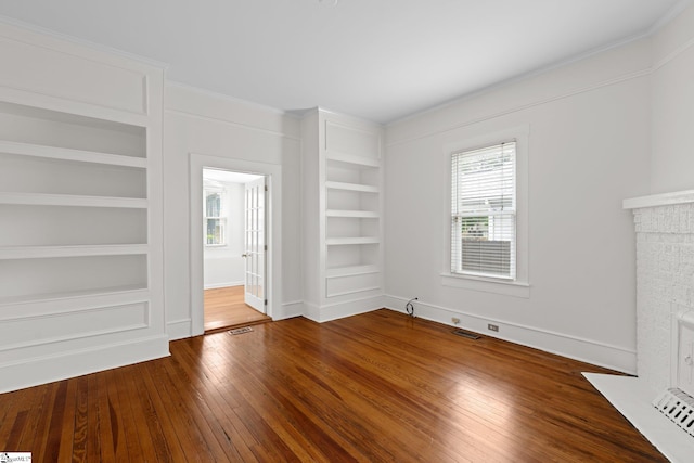 empty room with wood-type flooring, a brick fireplace, ornamental molding, and built in shelves