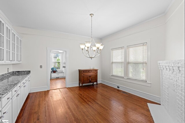 unfurnished dining area with a brick fireplace, ornamental molding, a notable chandelier, and dark hardwood / wood-style flooring