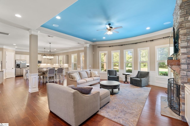 living room featuring ceiling fan with notable chandelier, dark hardwood / wood-style flooring, decorative columns, a stone fireplace, and ornamental molding