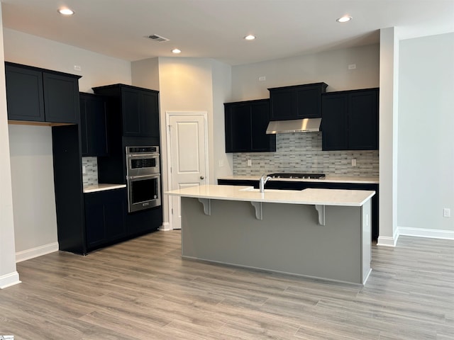 kitchen with backsplash, an island with sink, light hardwood / wood-style floors, a kitchen bar, and stainless steel double oven