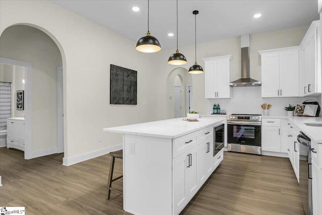 kitchen featuring wood-type flooring, hanging light fixtures, white cabinetry, stainless steel appliances, and wall chimney exhaust hood