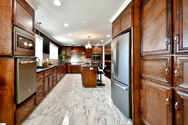 kitchen featuring hanging light fixtures, stainless steel appliances, custom range hood, decorative backsplash, and a center island with sink