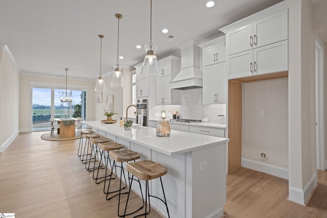 kitchen with custom range hood, a kitchen island with sink, decorative light fixtures, light hardwood / wood-style floors, and white cabinetry