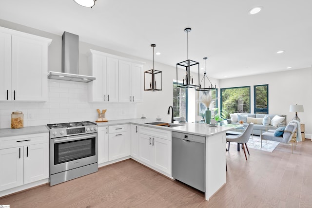 kitchen featuring wall chimney exhaust hood, stainless steel appliances, sink, white cabinets, and hanging light fixtures