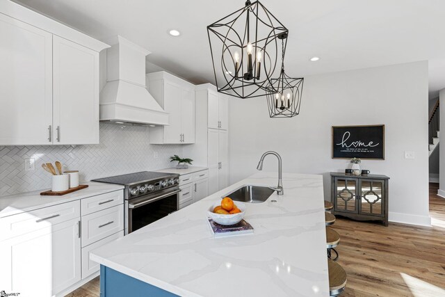 kitchen featuring custom exhaust hood, stainless steel electric stove, a kitchen island with sink, light hardwood / wood-style flooring, and sink