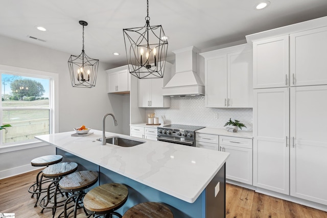 kitchen featuring sink, hanging light fixtures, stainless steel stove, premium range hood, and a center island with sink