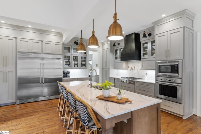 kitchen featuring custom exhaust hood, built in appliances, hanging light fixtures, and an island with sink