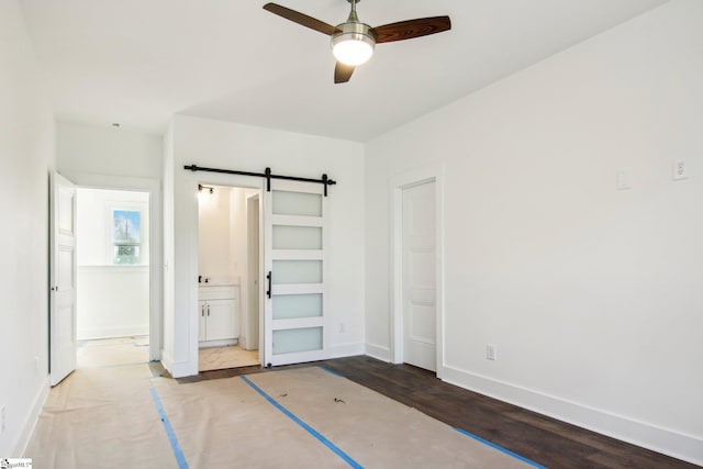 unfurnished bedroom featuring connected bathroom, a closet, wood-type flooring, a barn door, and ceiling fan