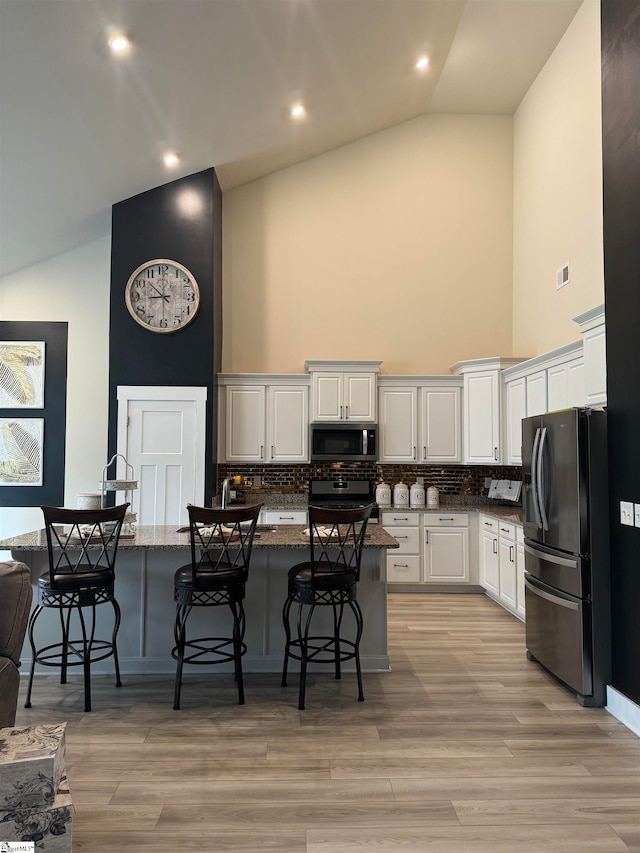 kitchen featuring a kitchen bar, white cabinetry, stainless steel appliances, dark stone countertops, and high vaulted ceiling