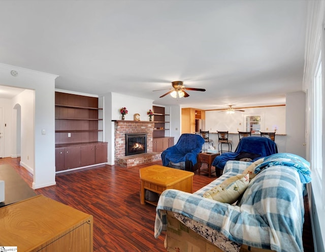 living room with built in shelves, ceiling fan, crown molding, a brick fireplace, and dark hardwood / wood-style flooring