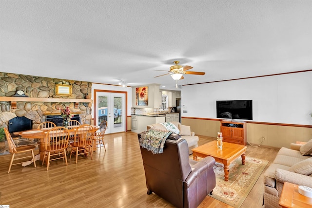 living room featuring ceiling fan, a textured ceiling, a stone fireplace, light hardwood / wood-style flooring, and french doors