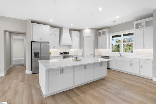 kitchen featuring appliances with stainless steel finishes, premium range hood, light wood-type flooring, a kitchen island, and pendant lighting